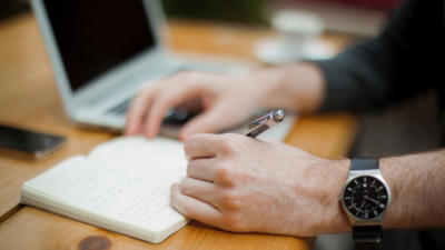 Close-up of man's hand holding pen and writing in journal at desk, other hand resting on adjacent computer keyboard, with watch on wrist and phone on desk in background